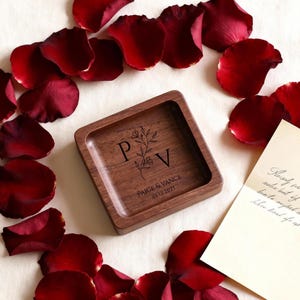 A square wooden ring dish engraved with the initials P and V and a date rests on a light fabric surrounded by scattered red rose petals and a handwritten note.