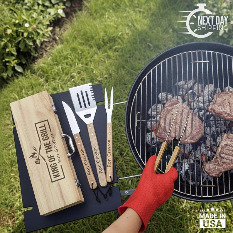 May include: A wooden storage box with the text KING OF THE GRILL and Ron Grayton engraved on the lid, accompanied by a matching knife, spatula, and fork with wooden handles. A red heat-resistant glove holds a metal fork over a charcoal grill.