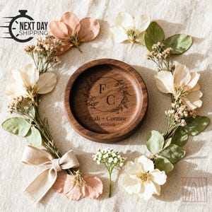 A round wooden ring dish with engraved initials F C sits on a fabric background decorated with dried flowers, green leaves, and a beige ribbon bow.