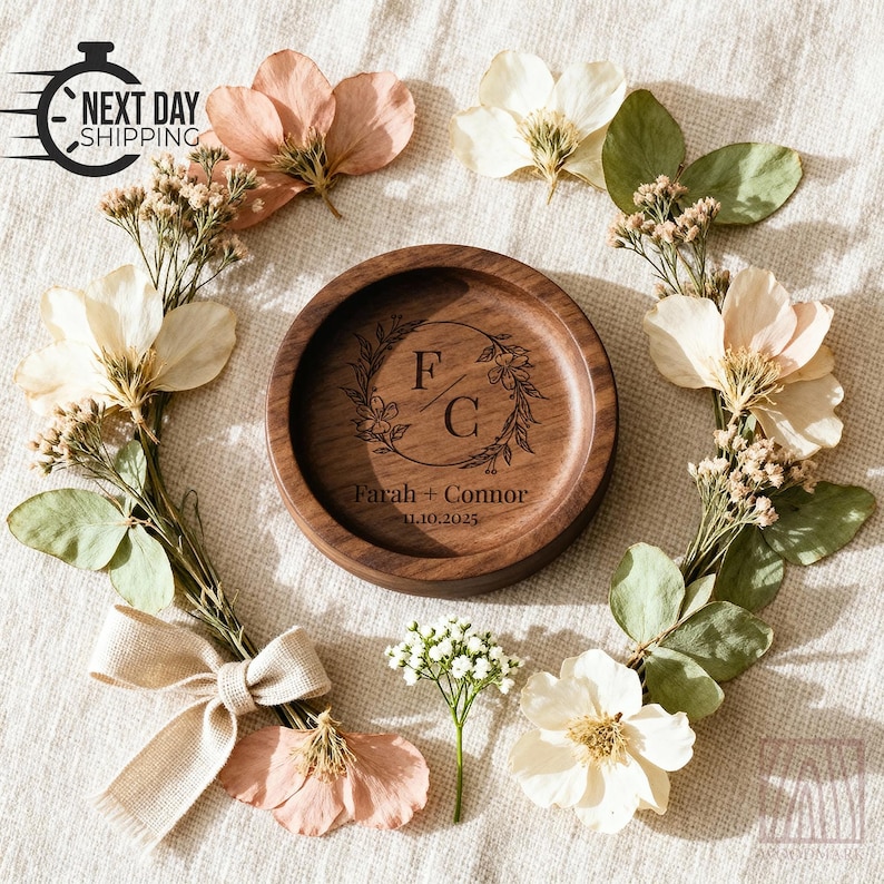 A round wooden ring dish with engraved initials F C sits on a fabric background decorated with dried flowers, green leaves, and a beige ribbon bow.
