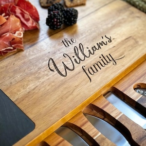 Op de afbeelding: Houten serveerplank met de gegraveerde tekst "the William's family". Het heeft een compartiment met kaasmesjes. De plank is gevuld met voedsel, waaronder prosciutto, bramen en druiven.