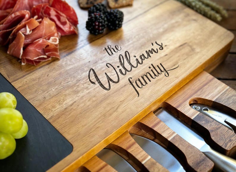 Op de afbeelding: Houten serveerplank met de gegraveerde tekst "the William's family". Het heeft een compartiment met kaasmesjes. De plank is gevuld met voedsel, waaronder prosciutto, bramen en druiven.