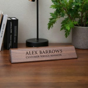 K&ouml;nnte beinhalten: Ein dunkelbraunes Namensschild aus Holz mit dem eingravierten Text "ALEX BARROWS" und "CUSTOMER SERVICE MANAGER". Das Namensschild befindet sich auf einem Holzschreibtisch. Eine kleine Pflanze und B&uuml;cher sind im Hintergrund.
