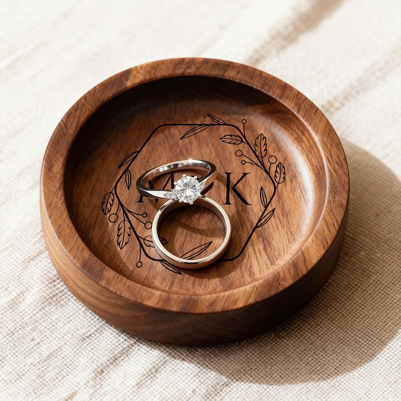 A round wooden ring dish engraved with a floral hexagonal frame and initials holds a diamond engagement ring and a plain band on a textured cloth.