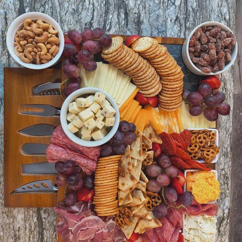 Op de afbeelding: Een houten charcuterieplank met een assortiment etenswaren. Bevat crackers, kaas, druiven, noten, salami en pretzels. De plank heeft ook een set kaasmessen. Het eten is gerangschikt op een houten plank met een donkere achtergrond.