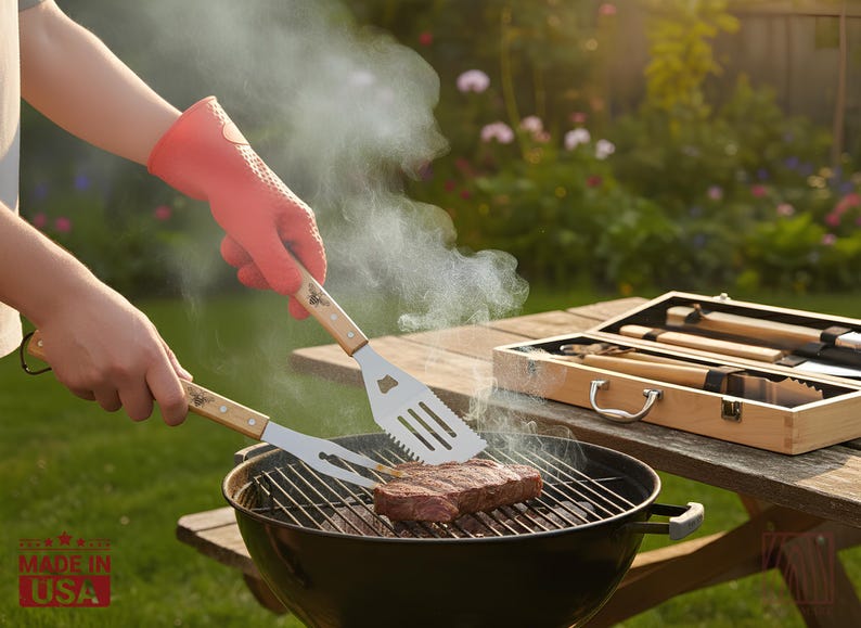 May include: A stainless steel spatula and a two-pronged fork with light wood handles, each featuring a small bee icon, are used to grill a raw steak on a black charcoal grill. A wooden carrying case with additional grilling tools sits nearby.