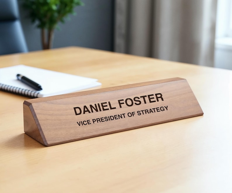 K&ouml;nnte beinhalten: Ein hellbraunes Namensschild aus Holz mit dem eingravierten Namen "DANIEL FOSTER" und dem Titel "VICE PRESIDENT OF STRATEGY" in Schwarz. Das Namensschild liegt auf einem hellen Holzschreibtisch.