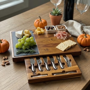 Op de afbeelding: Houten charcuterieplank met een leisteen kaasgedeelte, druiven, kaas, crackers en een selectie van gastronomische gerechten. Inclusief een lade met kaasmesjes en vorken. Wijnglazen, een fles wijn en pompoenen completeren het herfstthema.