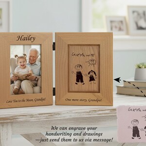 Op de afbeelding: Een houten fotolijst met twee panelen. E&eacute;n kant heeft een foto van een meisje en een man, met de naam "Hailey" erboven. De andere kant heeft een tekening van een persoon en een kind met de tekst "Opa en ik".