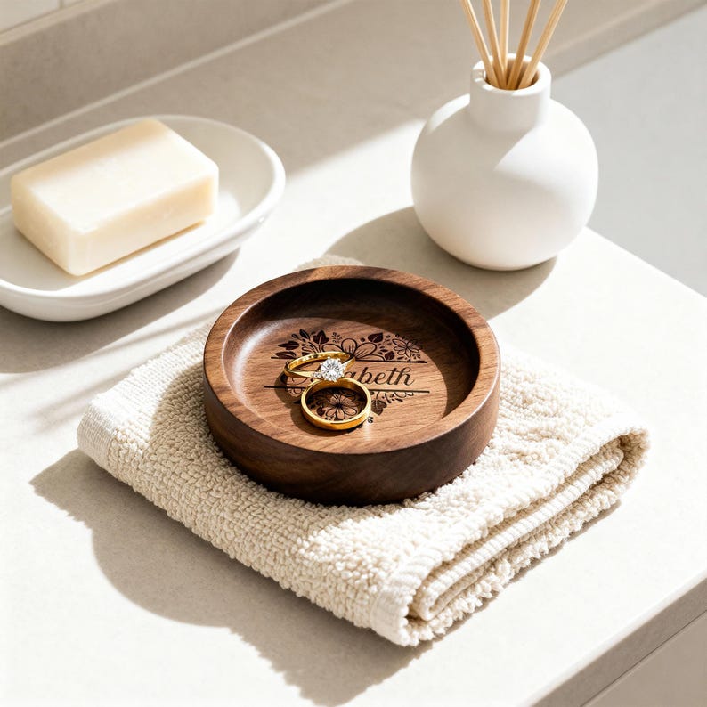 A round wooden ring dish sits on a folded cream hand towel holding two gold rings, positioned on a counter near a soap dish and diffuser.