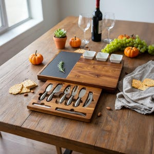 Op de afbeelding: Een houten kaasplankenset met een leistenen inzet, kleine witte kommen en een set kaasmesjes. De set bevat een fles wijn, wijnglazen en decoratieve pompoenen. De set staat op een houten tafel.