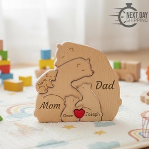 May include: Wooden bear family puzzle with the words "Mom," "Dad," and children's names. The puzzle features a red heart and is surrounded by other wooden toys. The image also includes the text "Next Day Shipping."