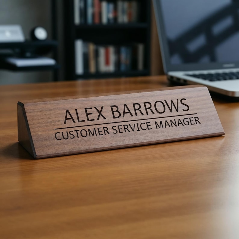 K&ouml;nnte beinhalten: Ein dunkelbraunes Namensschild aus Holz mit dem Namen "ALEX BARROWS" und der Berufsbezeichnung "CUSTOMER SERVICE MANAGER" in schwarzer Schrift. Das Schild hat eine dreieckige Form und liegt auf einem Holzschreibtisch.