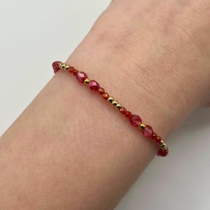 May include: A delicate bracelet featuring small, faceted red beads interspersed with gold-colored beads. The bracelet is strung on a thin, flexible cord, creating a simple yet elegant design. The bracelet is shown on a person's wrist.