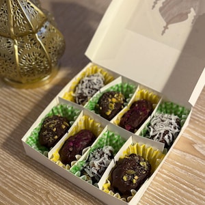 May include: A white box with a clear lid, filled with chocolate-covered dates. Some dates are topped with coconut flakes or pistachios. The box sits on a wooden surface, next to a gold lantern.
