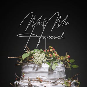 Puede incluir: Una tarta de boda blanca de tres pisos con glaseado texturizado, adornada con vegetación, bayas y ramitas. La tarta está coronada con un adorno blanco que dice "Mr & Mrs Hancock". La tarta se encuentra sobre un fondo oscuro.