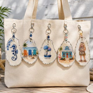 May include: A beige canvas tote bag featuring five hanging oyster shell ornaments. The ornaments display painted designs of a blue seahorse, beach chairs, blue hydrangeas with the text CAPE COD, a beach house, and a person holding a sign.
