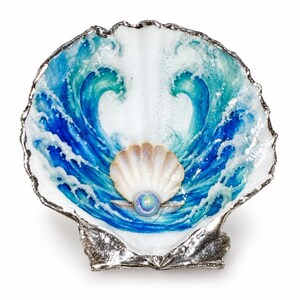 May include: A large scallop shell dish with a silver-toned metallic rim. The interior features a hand-painted design of two blue and teal ocean waves crashing toward the center, where a smaller shell holds a single iridescent spherical pearl.