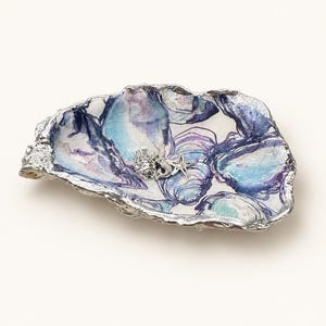 Può includere: Un piatto decorativo a forma di guscio d'ostrica con un interno in decoupage caratterizzato da un motivo astratto blu e viola. I bordi sono rifiniti con una bordatura in argento metallizzato. All'interno ci sono tre piccoli ciondoli argentati: un pesce, un cavalluccio marino e una stella marina.