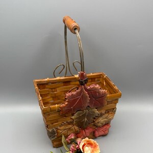 May include: A woven basket with a wooden handle and decorative metal accents. The basket is adorned with sculpted leaves and a cluster of artificial flowers in shades of peach and pink. The basket is approximately 7 inches tall.