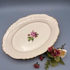 May include: An oval-shaped, off-white serving platter with a gold trim and a pink rose floral design in the center. The platter has a scalloped edge. Artificial flowers are arranged near the platter.