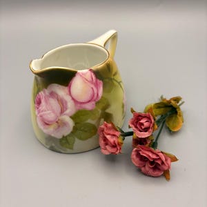 May include: A small, white ceramic creamer with a handle and a gold rim. The creamer is decorated with pink roses and green leaves. A sprig of pink roses rests beside the creamer.