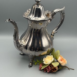 May include: A silver-colored teapot with ornate detailing, including a decorative handle and spout. The teapot has a lid with a finial and rests on three small feet. A small bouquet of flowers in shades of yellow, peach, and burgundy is next to the teapot.