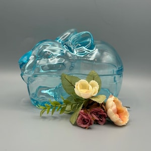 May include: A light blue glass bunny-shaped trinket box with a lid. The box is decorated with a small bouquet of artificial flowers in shades of cream, peach, and burgundy. The bunny has long ears and a detailed face.