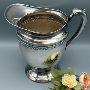 May include: A silver-colored pitcher with a decorative handle and a wide spout. The pitcher has a detailed rope-like design around the middle and sits on a pedestal base. A small floral arrangement with yellow and pink roses is in the foreground.