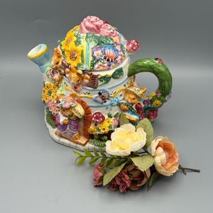 May include: A decorative ceramic teapot with a whimsical garden theme. The teapot features colorful flowers, mushrooms, and small animal figurines. The handle is green with a ladybug, and a small bouquet of flowers rests nearby.