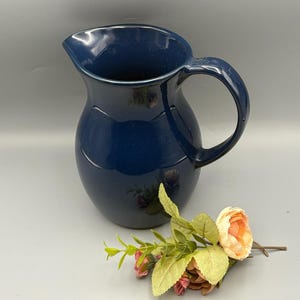 May include: A deep navy blue ceramic pitcher with a smooth, glossy finish, a curved handle, and a wide pouring spout. Beside the base of the pitcher lies a small decorative sprig featuring light peach-colored flowers and green leaves.