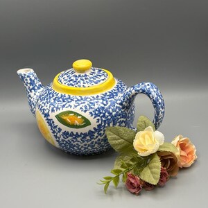 May include: A ceramic teapot with a blue and white floral pattern and yellow accents. The teapot features a lemon design and a matching lid. A small bouquet of roses in shades of pink, peach, and yellow rests beside the teapot.