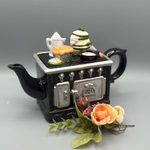 May include: A black and silver teapot shaped like a vintage stove, with miniature food and kitchenware on top. The teapot has a handle and spout, and is decorated with a small floral arrangement.