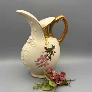 May include: Cream-colored ceramic pitcher with a gold handle and rim. The pitcher features embossed floral designs and hand-painted pink flowers. A small bouquet of pink flowers rests near the pitcher.