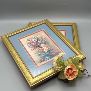 May include: Two framed prints with gold-colored frames. The prints feature a floral arrangement with a cherub. The frames are rectangular with a gold-colored trim. A small floral arrangement with a peach-colored rose is placed on top of the frames.