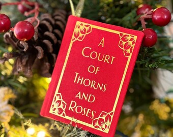 May include: A red Christmas tree ornament shaped like a book with gold trim and the title "A Court of Thorns and Roses". The ornament is hanging on a Christmas tree with red berries and pinecones.