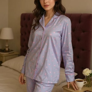 May include: A light purple pajama set with a pink heart pattern. The long-sleeved top and pants have pink trim. The pajamas are button-down and are made of a soft material. The model is standing in a bedroom.