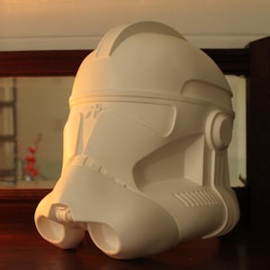 May include: A white, molded plastic helmet with a distinctive design, featuring a rounded top, visor, and side details. The helmet has a smooth, matte finish and is likely a replica or prop from a science fiction franchise.
