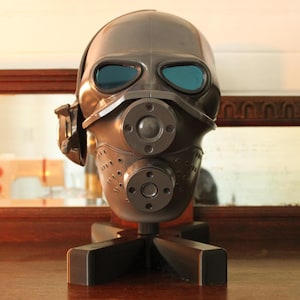 May include: A dark gray gas mask with blue tinted lenses and circular attachments. The mask is mounted on a black stand, and is displayed on a wooden surface. The background includes a mirror and a glimpse of other objects.