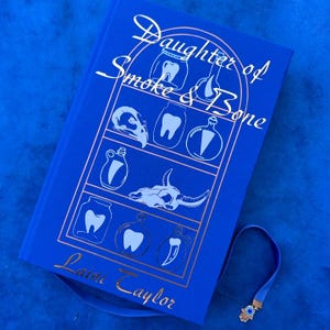 May include: A blue hardback book titled "Daughter of Smoke & Bone" with gold foil detailing. The cover displays illustrations of teeth, bottles, and a skull. A blue bookmark with a hand charm is attached.