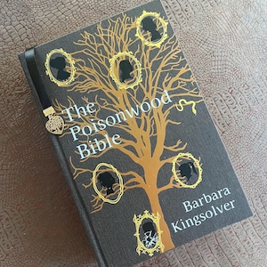 May include: A hardcover book titled The Poisonwood Bible by Barbara Kingsolver with a black cover featuring a gold tree illustration, five gold-framed silhouettes, and a black ribbon bookmark with a gold heart-shaped tree charm attached.