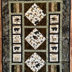 May include: A quilted blanket with a nature theme. The quilt features a dark green and brown color scheme with images of bears, bison, and other wildlife. The quilt has a diamond pattern in the center and rectangular panels with animal illustrations.