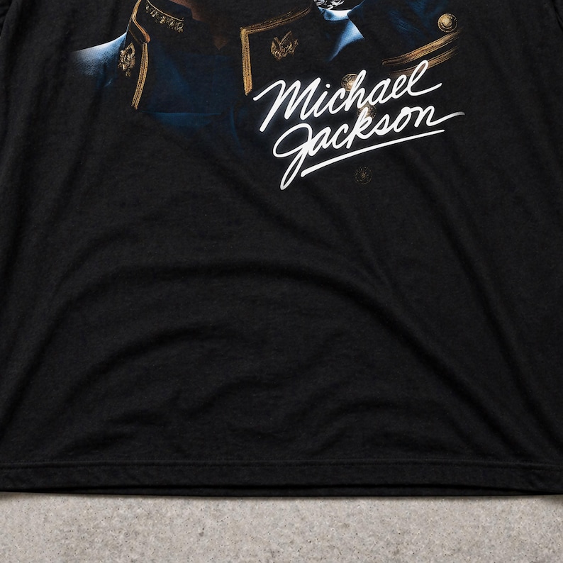 May include: Black t-shirt featuring the name "Michael Jackson" in white script. The shirt has a close-up image of a person in a military-style jacket. The shirt is made of a soft, dark fabric.