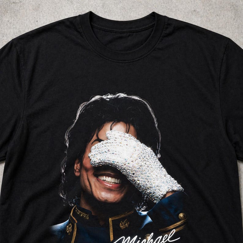 May include: Black t-shirt featuring a portrait of a smiling person wearing a sequined glove, covering their eyes. The person has dark hair and is wearing a blue and gold jacket. The name "Michael" is written in cursive.