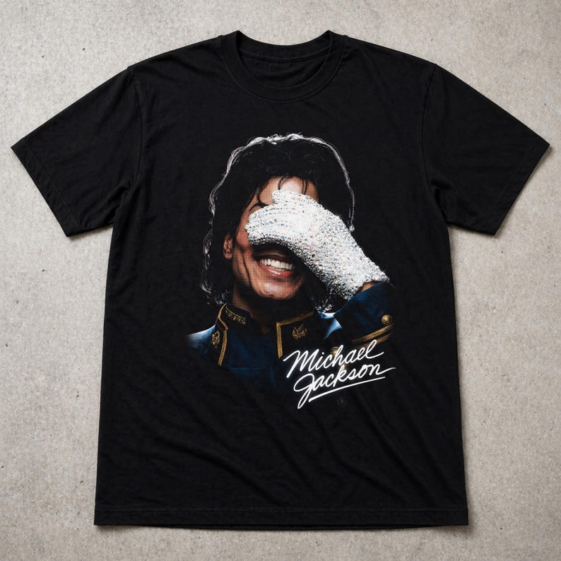 May include: Black t-shirt featuring a portrait of Michael Jackson. The image shows Jackson smiling, with his face partially covered by a white, jeweled glove. The words "Michael Jackson" are written in cursive below the portrait.