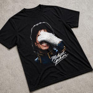 May include: Black t-shirt featuring a graphic of Michael Jackson smiling, with his hand covering his eyes and wearing a white glove. The text "Michael Jackson" is written in white script.