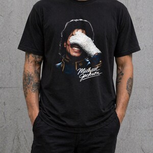 May include: Black t-shirt featuring a smiling portrait of Michael Jackson, with his face partially covered by a white glove. The image includes the text "Michael Jackson" in a cursive font, capturing a vintage aesthetic.