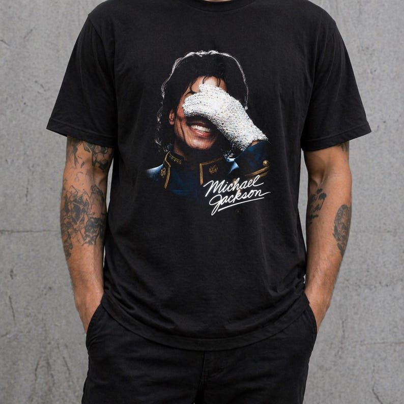 May include: Black t-shirt featuring a smiling portrait of Michael Jackson, with his face partially covered by a white glove. The image includes the text "Michael Jackson" in a cursive font, capturing a vintage aesthetic.