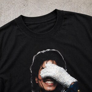 May include: Black t-shirt featuring a smiling portrait of a person wearing a sequined glove over their eyes. The image is a close-up of the person's face, with the glove and hair in sharp focus. The shirt's neckline is visible.