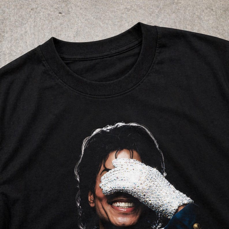 May include: Black t-shirt featuring a smiling portrait of a person wearing a sequined glove over their eyes. The image is a close-up of the person's face, with the glove and hair in sharp focus. The shirt's neckline is visible.